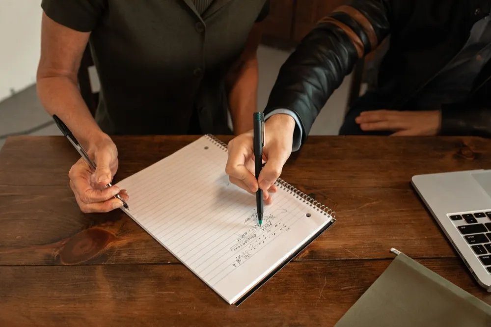 Two people writing notes together at a wooden table.